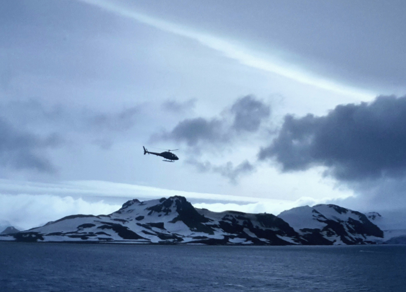 Helicóptero sobrevoando montanhas cobertas de neve na Península Antártica durante expedição brasileira